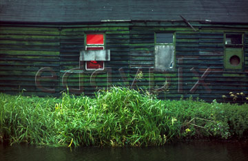 Comp image : dere0109 : Dark, derelict old riverside building with a red painted boarded up window and green grass on the river bank