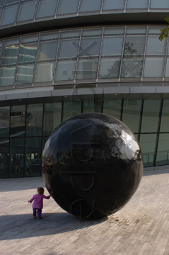 Comp image : lond010046 : 'Black ball' sculpture outside London's City Hall, dwarfing a tiny girl who is trying to move it