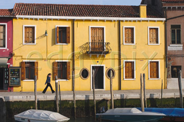 Comp image : ven021407 : Girl striding past a bright warm yellow building with shuttered windows in late afternooon sun in Murano, Venice, Italy; boats moored on the canal in front