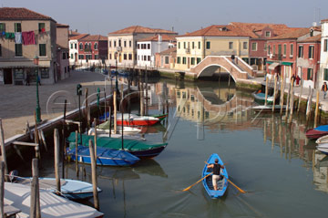 Comp image : ven021417 : A lone boatman rows a blue boat along a smooth canal in Murano, Venice, Italy, in evening sunshine; a perfect reflection of a bridge and buildings in the background