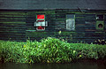 Dark, derelict old riverside building with a red painted boarded up window and green grass on the river bank