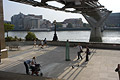 Three small groups of people in the shadows under the Millennium Bridge over the River Thames in London, England, with a view across the river in the background