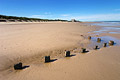 A line of wooden posts in sunshine on a deserted sandy beach at low tide on the flat North Norfolk coast of England, under a clear blue sky