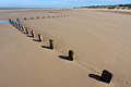 A zigzag line of wooden posts in sunshine on a deserted sandy beach at low tide on the flat North Norfolk coast of England, under a clear blue sky