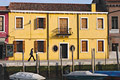 Girl striding past a bright warm yellow building with shuttered windows in late afternooon sun in Murano, Venice, Italy; boats moored on the canal in front