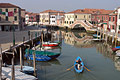 A lone boatman rows a blue boat along a smooth canal in Murano, Venice, Italy, in evening sunshine; a perfect reflection of a bridge and buildings in the background