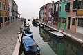 Looking along a perfect calm canal in Burano towards the Venice lagoon, with boats moored by the colourful houses and another entering the canal in the distance